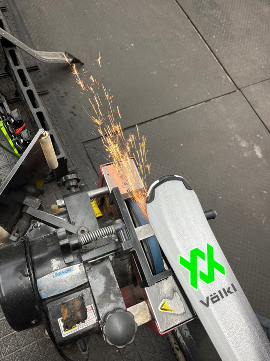 A Ski and Snowboard Service Manager sharpening Völkl ski edges on a tuning machine, generating bright orange sparks.