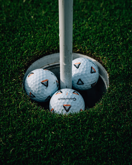 Close-up of premium dimpled golf balls with TaylorMade branding inside a hole on a professional grass green.