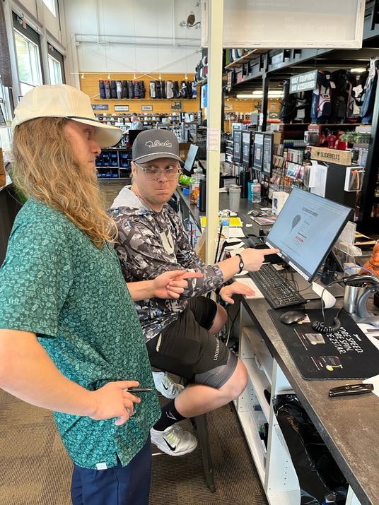 A retail manager providing backend system training to an associate at a pro shop counter, demonstrating leadership skills.