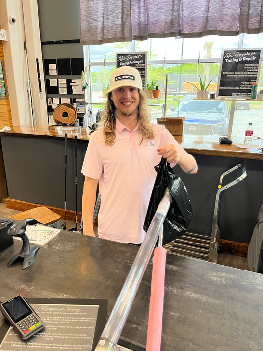 Professional retail sales associate at a pro shop counter featuring ski tuning and golf equipment service menus.